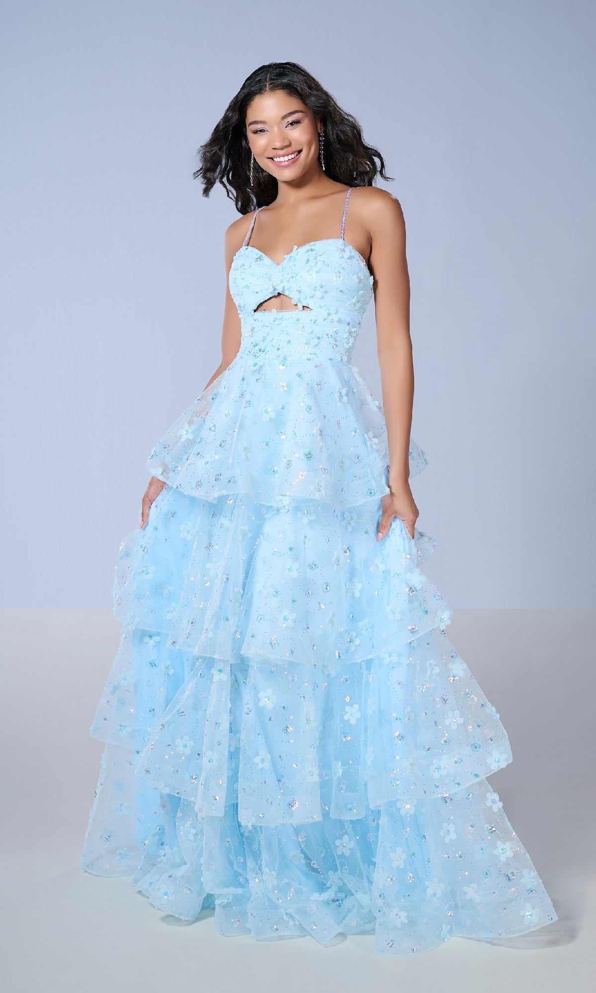 A woman wearing the Tiffany 16144 long prom dress, featuring a light blue sweetheart neckline, spaghetti straps, midriff cutout, and ruffled A-line skirt, stands against a plain background, smiling.